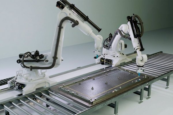 Two white Kuka robotic arms, mounted on a rail, move along a piece of carbon-reinforced concrete and install wires for the manufacture of the product.
