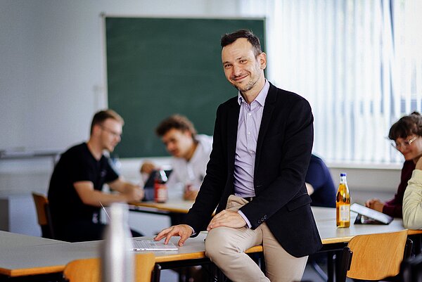 Volker Gruhne. (Foto: Anne Schwerin/HTWK Leipzig) Mann sitzt auf Tisch in Seminarraum, im Hintergrund Studierende